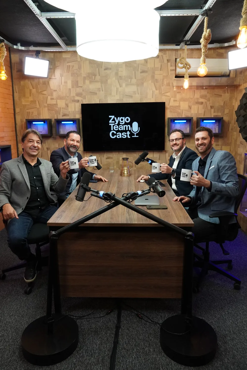 O estúdio do Zygo Team Cast com os apresentadores à mesa, segurando canecas do podcast