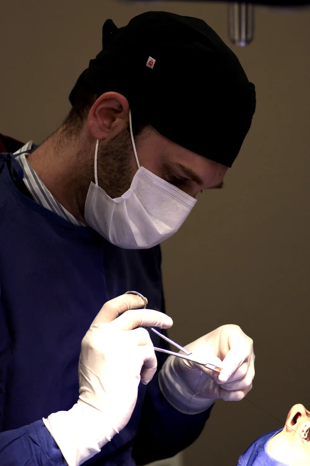 Dr. Guilherme Speck, de máscara e touca, realizando uma sutura com pinça e fio
